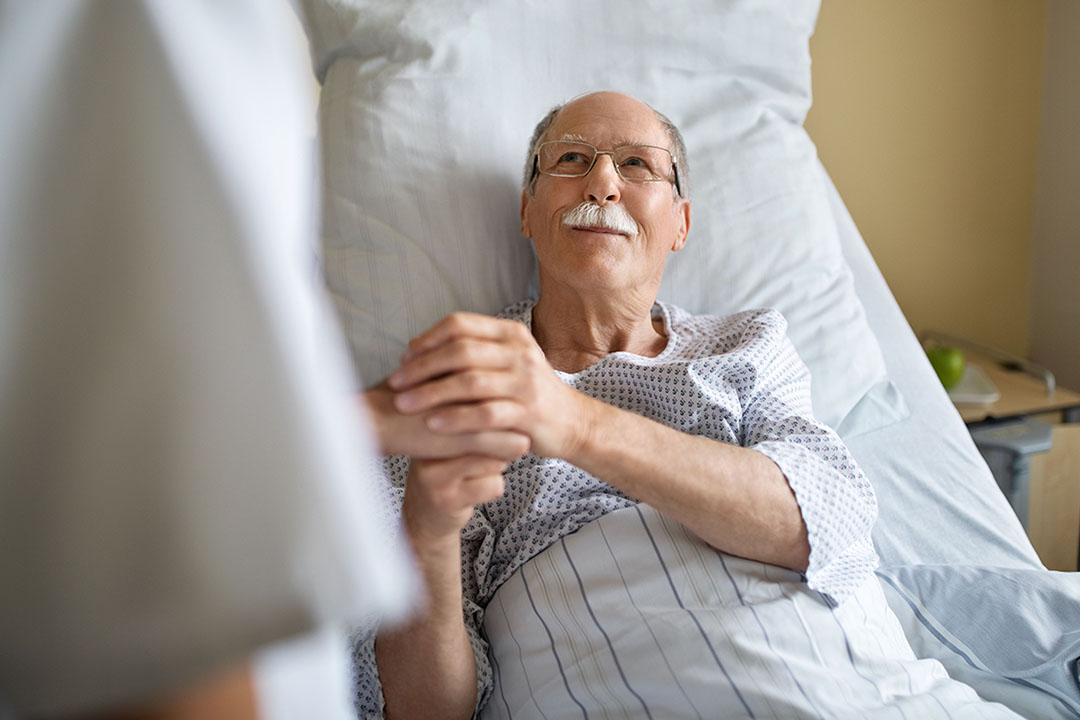 Ein älterer Mann liegt im Krankenbett und hält die Hand eines Arztes