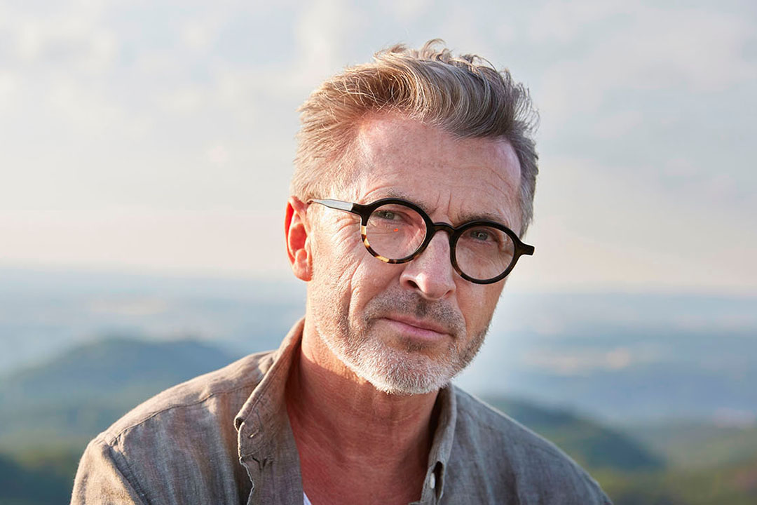 Älterer Mann mit Brille schaut in die Kamera, im Hintergrund ist eine Hügellandschaft zu sehen