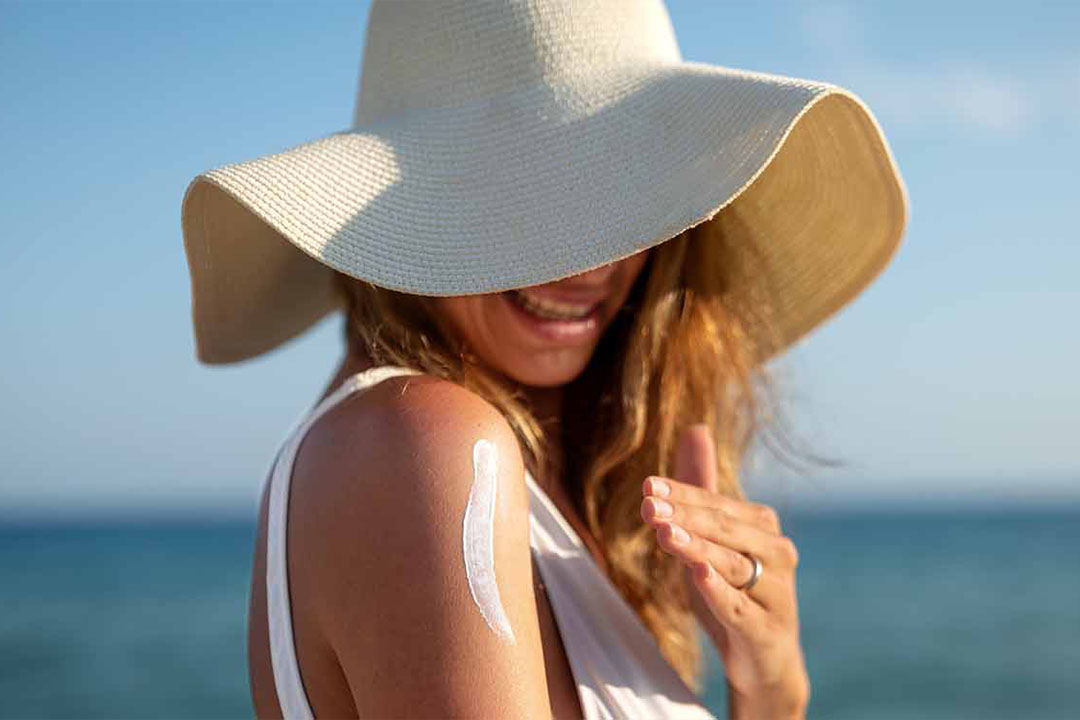 Frau am Strand die einen Hut trägt und sich die Schulter mit Sonnencreme eincremt