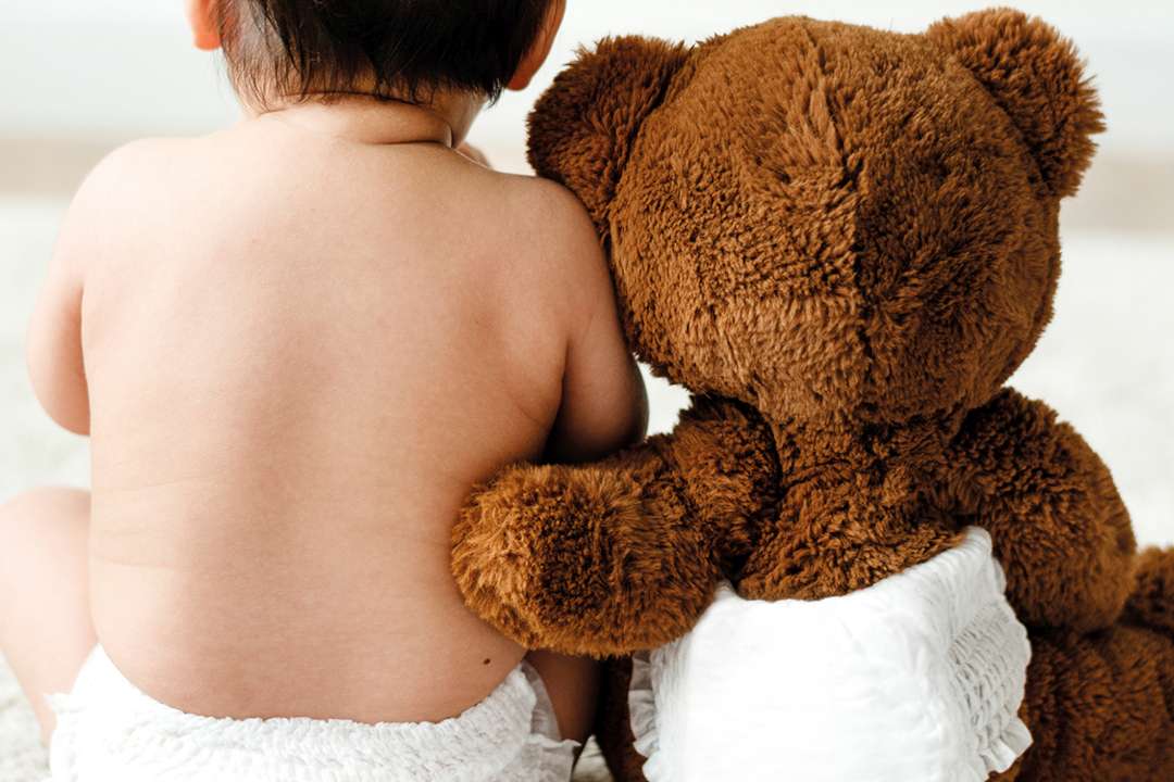 Ein Kleinkind sitzt von uns weg gedreht auf dem Boden und wird von einem Teddy umarmt.