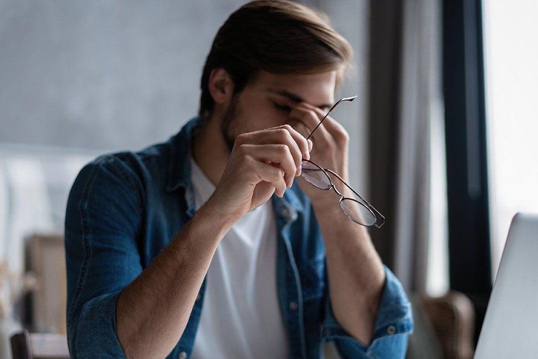 Ein Mann hält seine Brille in der Hand das er Schmerzen an der Nasenwurzel hat