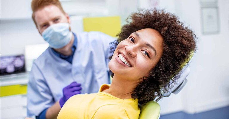 Junge Frau sitzt auf dem Behandlungsstuhl und lächelt mit strahlend weissen Zähnen. Der Zahnarzt sitzt im Hintergrund auf einem Hocker.