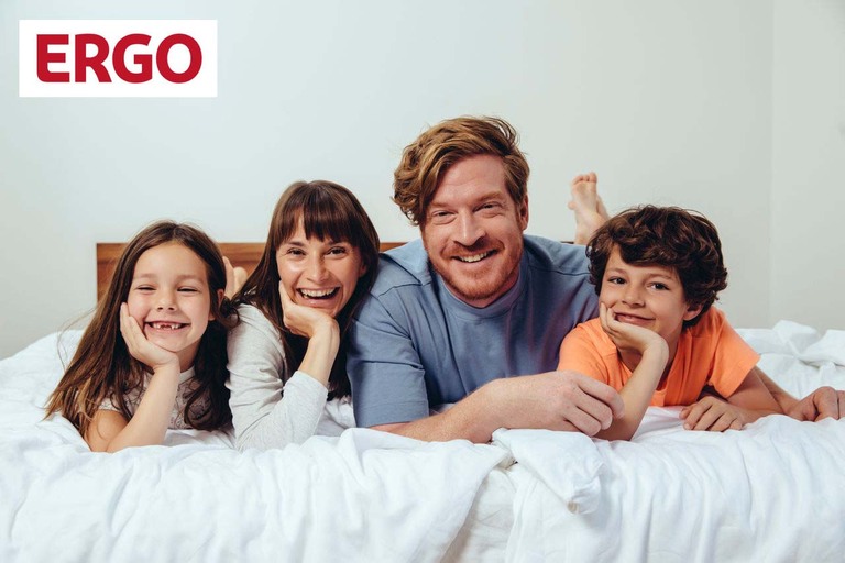 Eine Familie liegt auf dem Bett und lächelt in die Kamera Mit Logo der ERGO