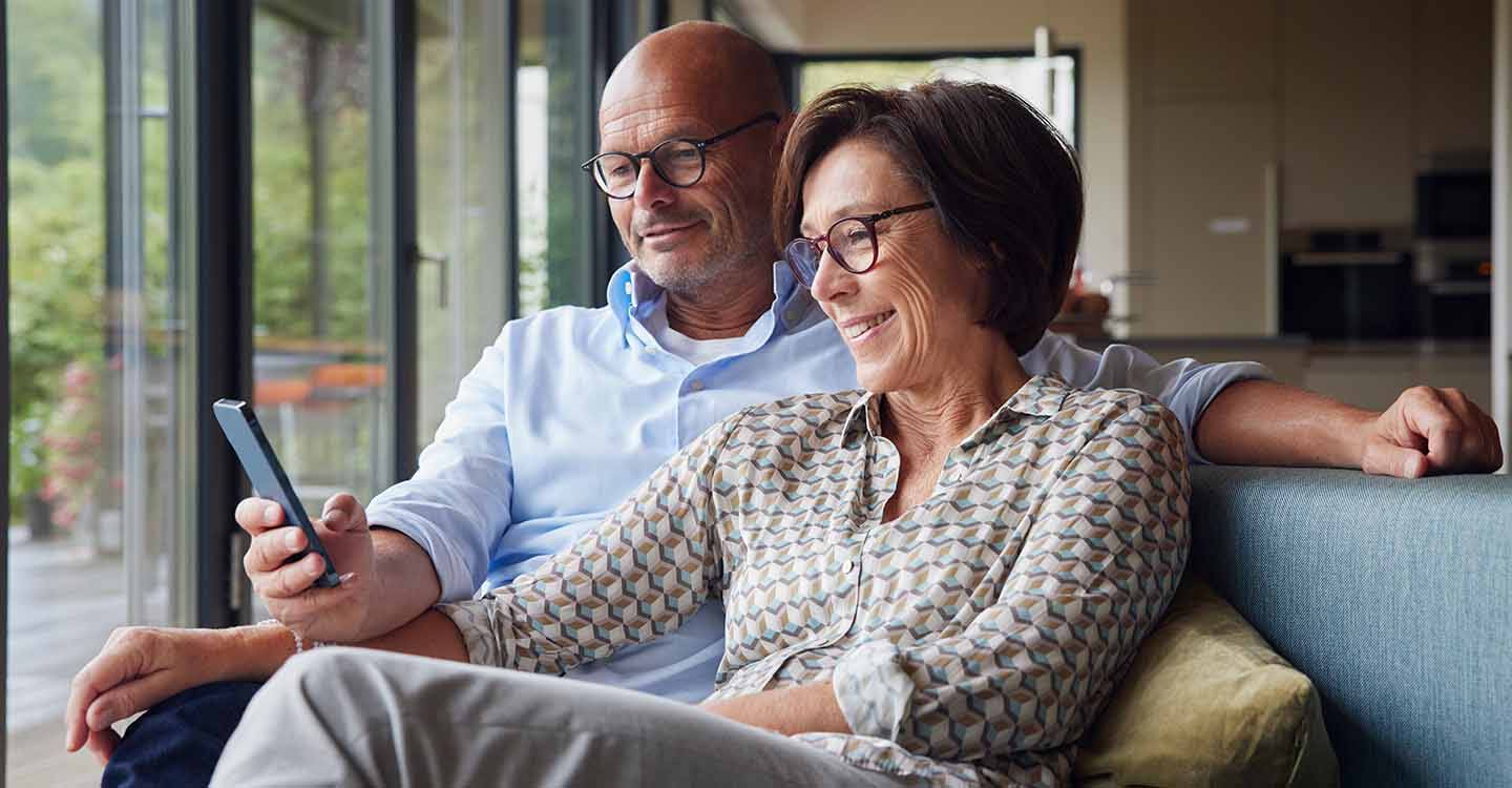 Glückliche Frau mit einem Mann, der sein Smartphone benutzt, während sie zu Hause auf dem Sofa sitzen