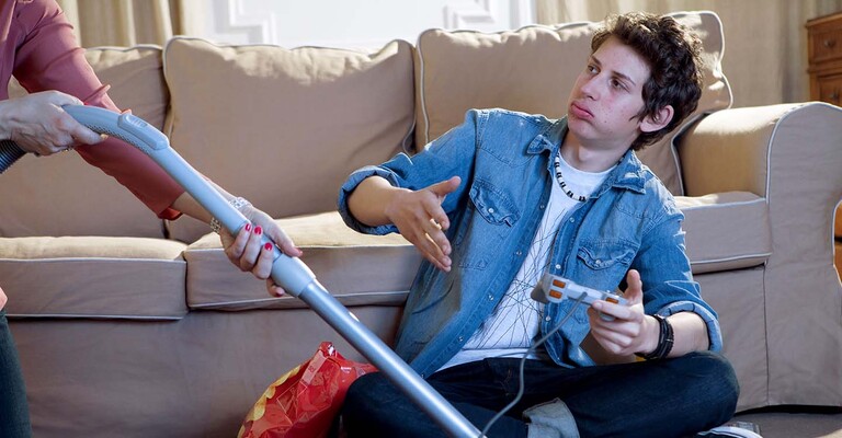 Junger Mann sitzt auf dem Teppich beim Gaming und Mutter staubsaugt um ihn herum