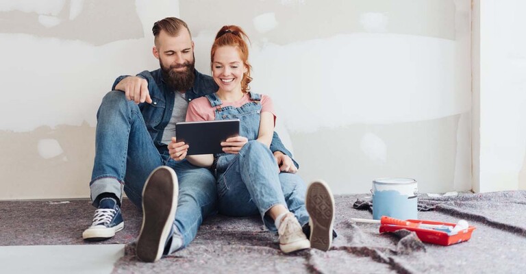 Junges Paar sitzt auf dem Boden und schaut auf ein Tablet. Der Raum wird gerade renoviert.