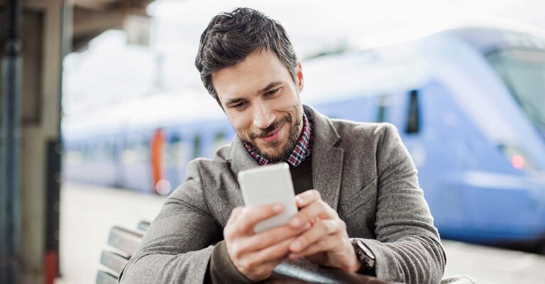 Ein Mann sitzt auf einer Bank am Bahngleis und schaut gespannt und freudig auf sein Handy, während er auf die Bahn wartet.