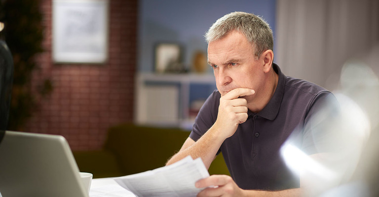 Mann sitzt mit Unterlagen vor einem Laptop und denkt nach