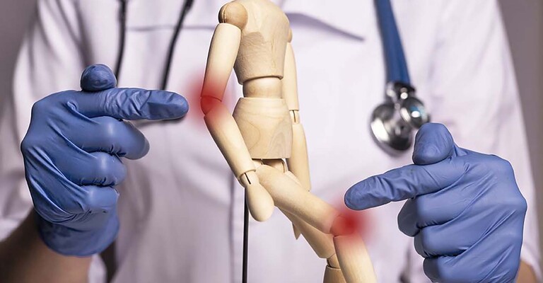 Zwei Hände in Gummihandschuhen zeigen auf die markierten Gelenke einer Holzpuppe