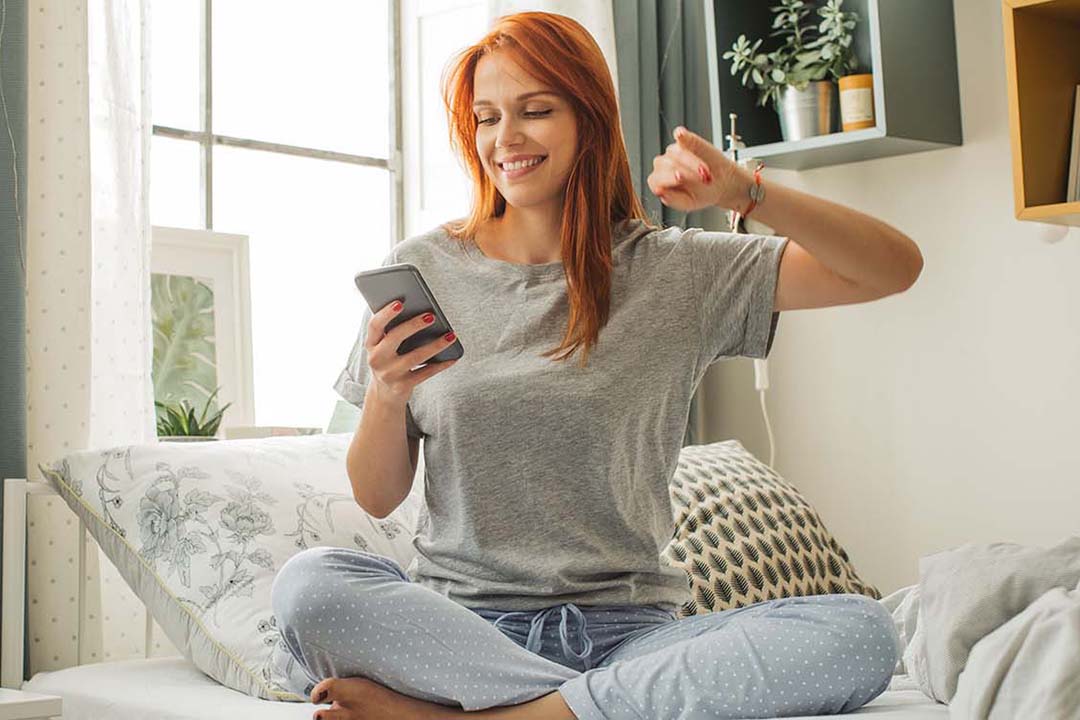 Eine Frau sitzt auf ihrem Bett und schaut strahlend auf ein Smartphone